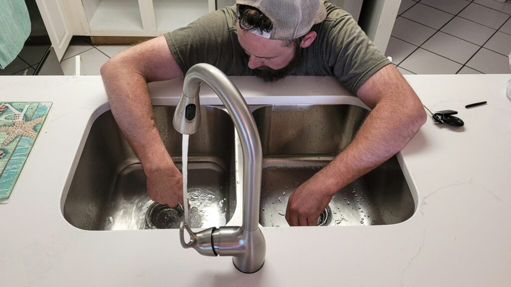 A man in a kitchen fixing a sink, with water flowing from the faucet.