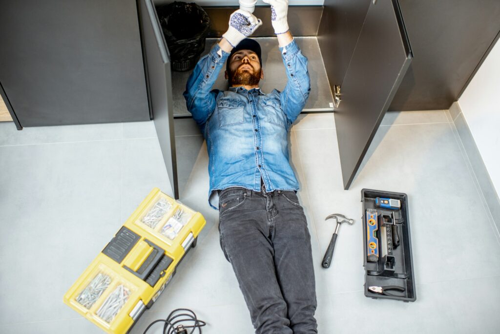 A man lies on the floor surrounded by plumbing tools and a toolbox, focused on a repair task.